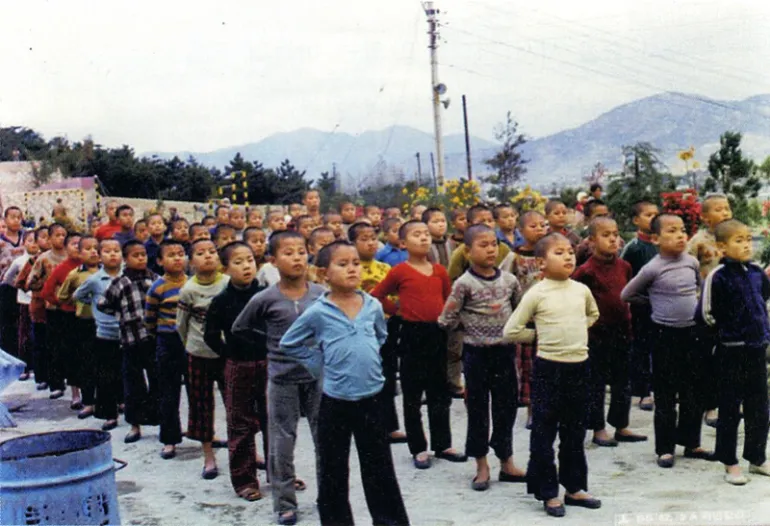 Children with shaven heads stand in queues, with hands behind their backs.