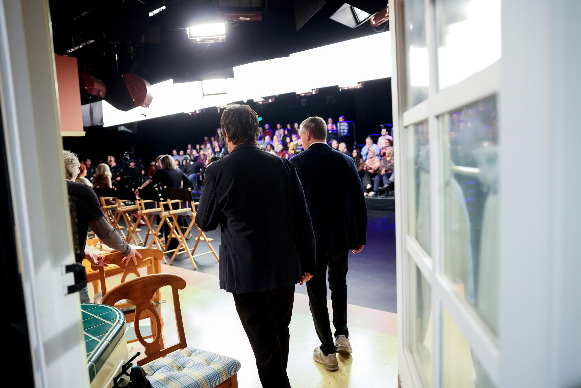 Two men walking toward the front of a stage where several directors chairs are lined up and audience sits before them.