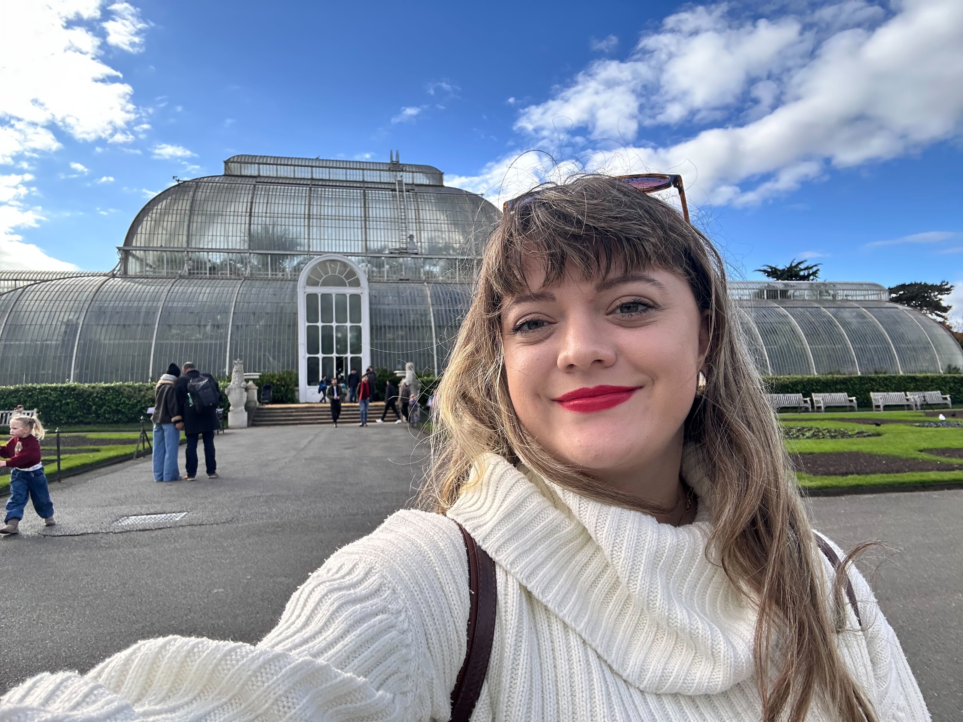 A woman with red lipstick smiling in front of the Palm House at Kew Gardens.