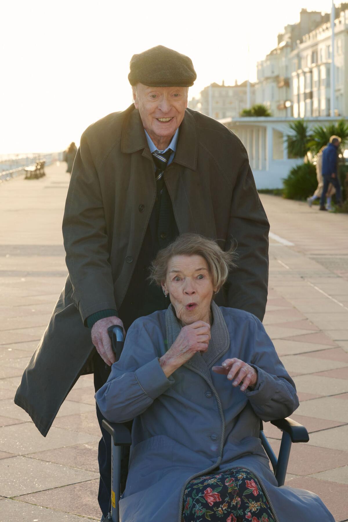 An elderly man in a hat and coat pushes a woman with a surprised expression in a wheelchair.