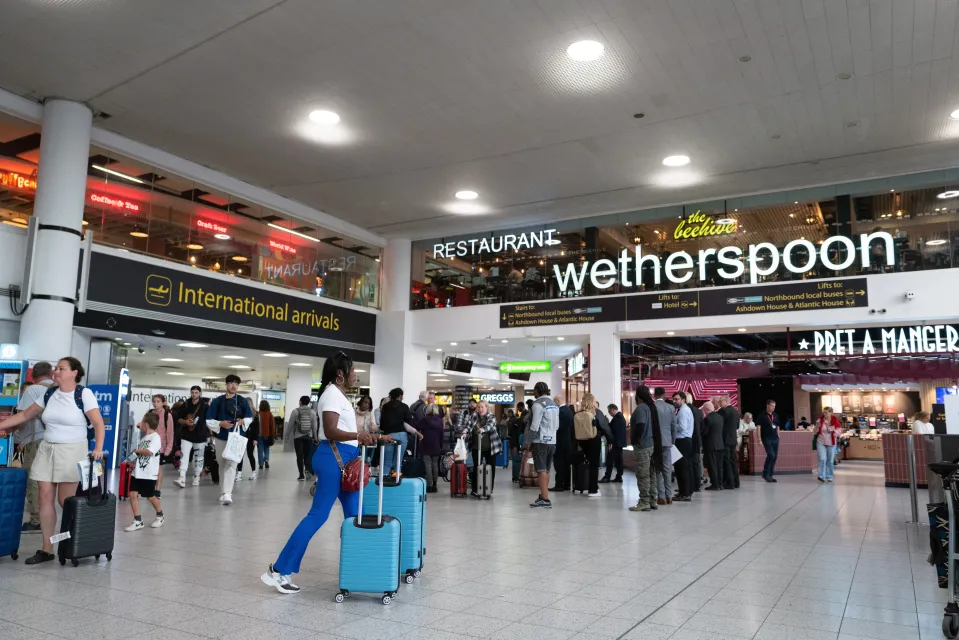 Travelers at Gatwick South Terminal in the international arrivals area, with signs for Wetherspoon and Pret A Manger visible.