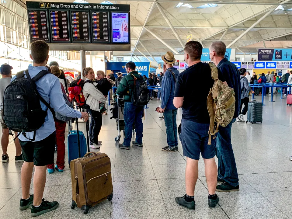 People and luggage at a check-in board displaying flight information and advertisements at Stansted Airport.