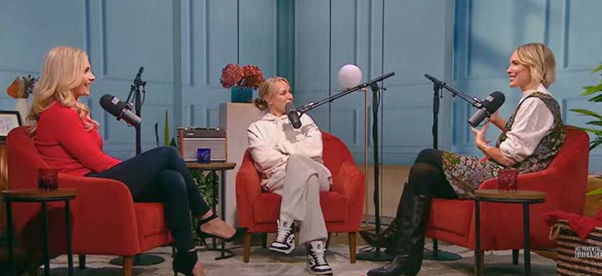 Two individuals are seated on red chairs in an indoor setting, engaged in an interview. Both are holding microphones positioned on stands before them. The setting includes a light blue wall, a small table with a potted plant to the right, and a side table with a cup to the left.