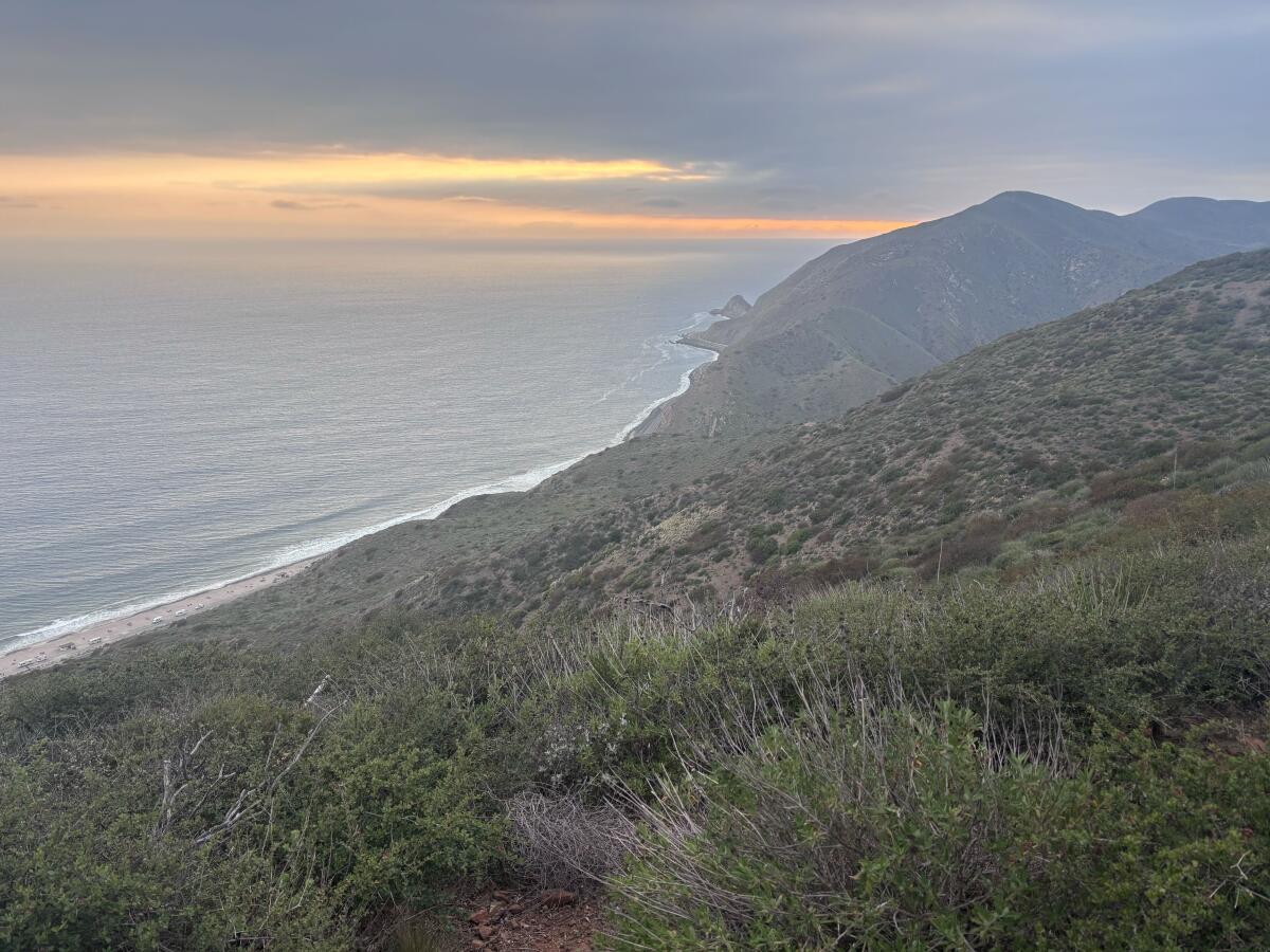 The coastline with splashing waves amid a pinkish orange sunset with dark blue clouds.
