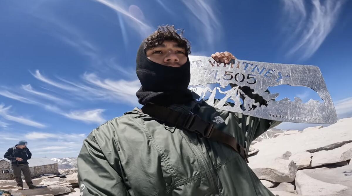 A hiker with a balaclava holds a metal summit sign at the top of Mt Whitney.