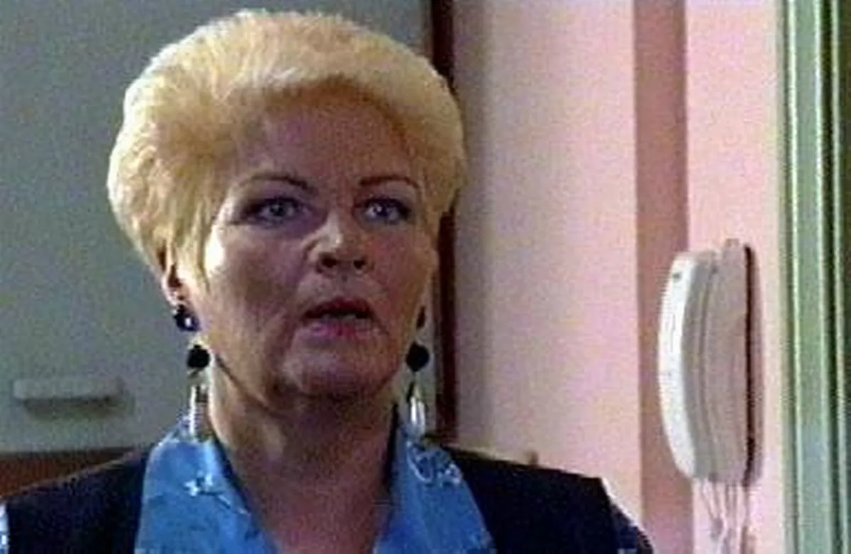 An adult female with short blonde hair and wearing a blue top is standing and appears to be speaking, positioned against a pink wall with a mounted intercom visible.