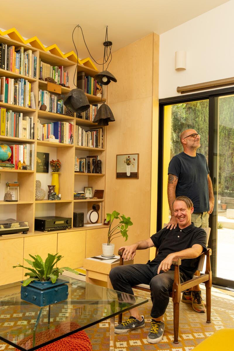 Two people, one sitting and one standing, near a large bookcase and a glass table.