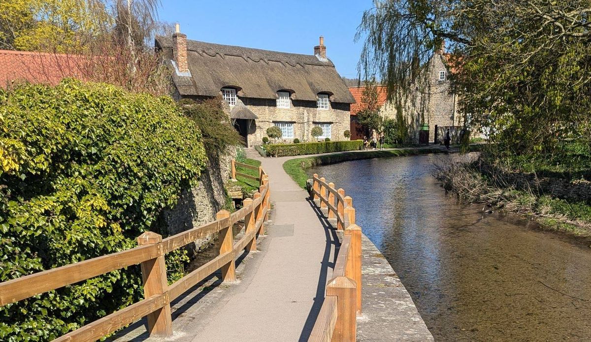 A serene scene featuring a wooden walkway along a tranquil river, bordered by neatly trimmed hedges and several charming cottages with thatched roofs. The pathway extends toward a row of houses, with trees and greenery framing the landscape.