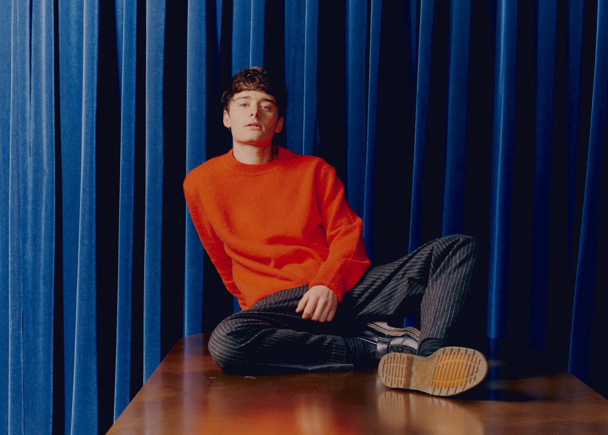 A guy in an orange sweaters sits atop a table 