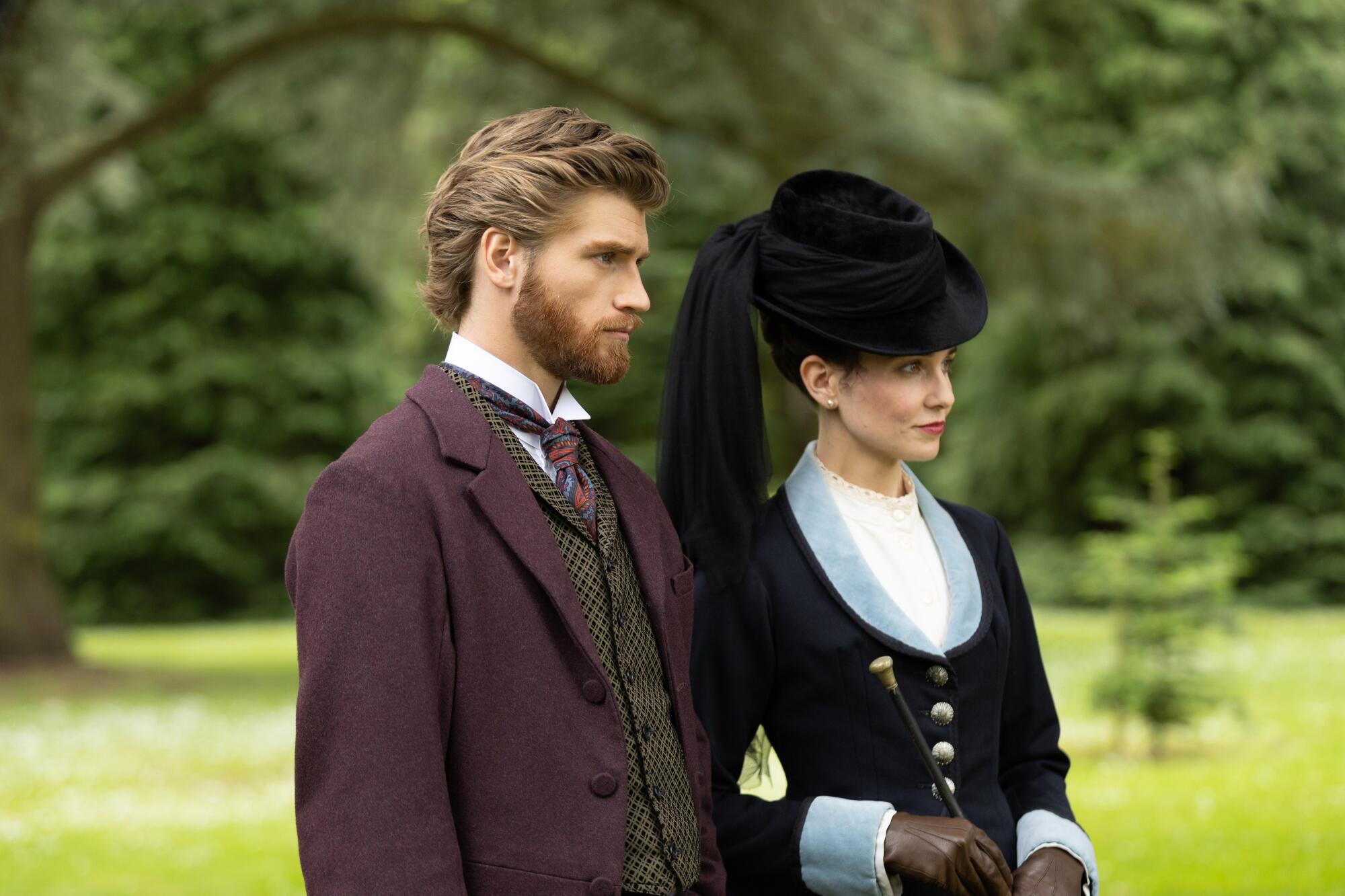 A man in a brown suit stands with a woman in a black and blue coat and black hat.