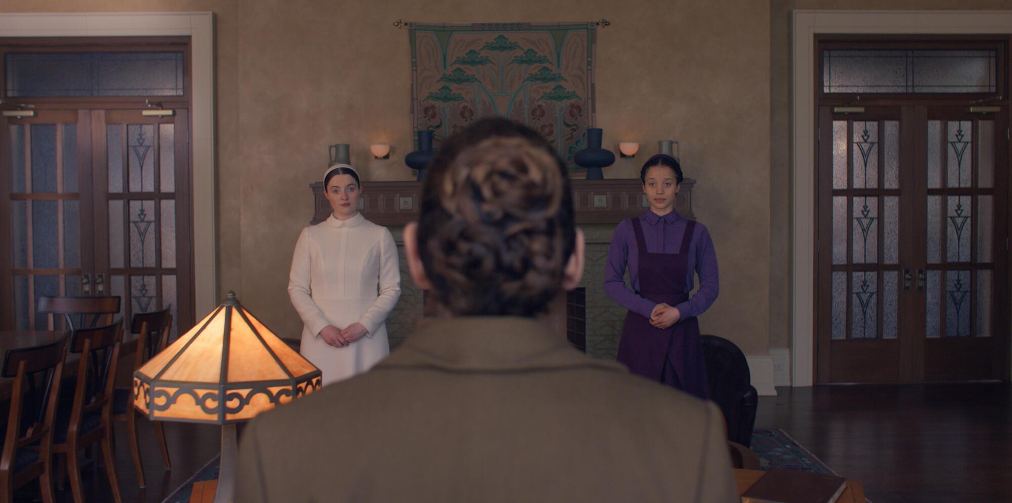 Two teenage girls stand on either side of a bed looking at a woman seen from behind.