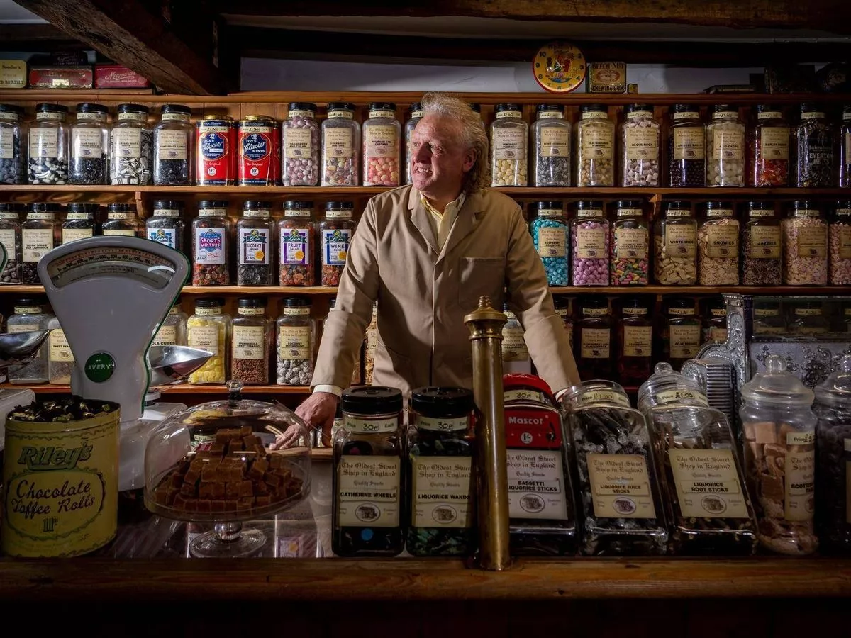 An individual, dressed in a suit, is standing behind a counter in a store. The counter is stocked with a variety of colorful jars and containers, some of which are labeled. The shelves behind the person are also filled with numerous jars and products, creating an organized and vibrant display.