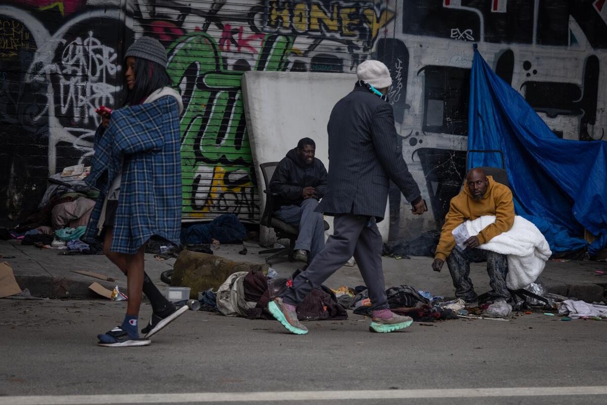 People walk by people lying or sitting on a sidewalk.