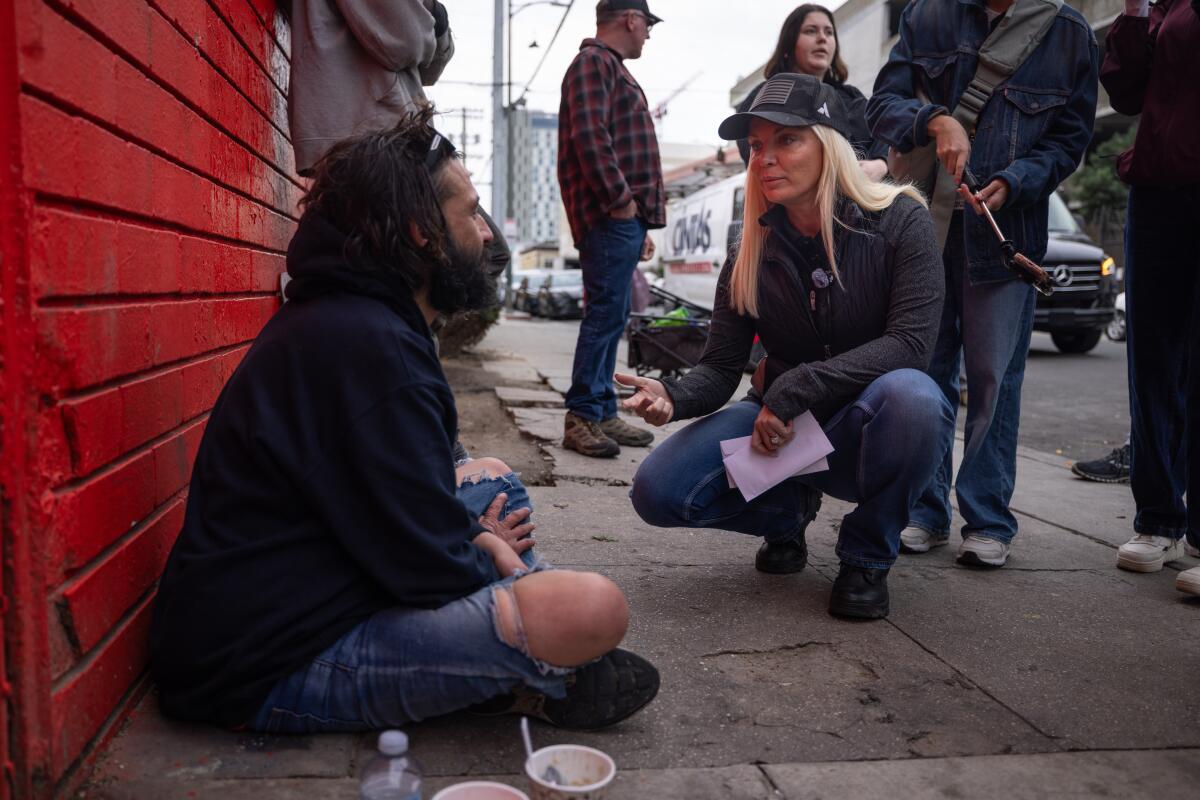 A woman crouches down to talk to a person sitting on a sidewalk.