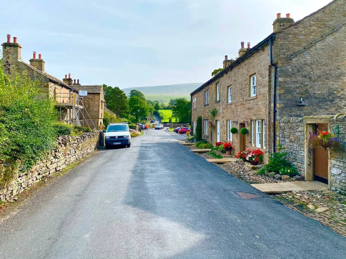 An image depicts a quiet street in a countryside village, featuring stone-built houses and greenery. A vehicle is parked on the side of the road, indicating a peaceful and serene rural setting.