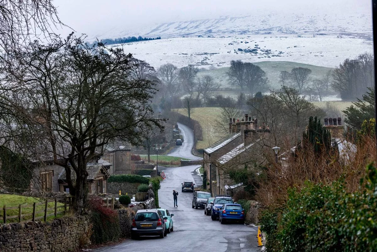 An aerial view of a street in a rural setting reveals a foggy landscape with hills and trees. Several cars are parked along the road, and a person is walking on the sidewalk. The scene is bathed in a soft, diffused light.
