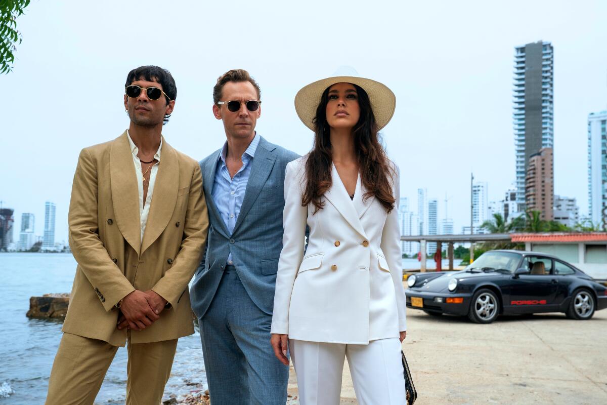 A man in a tan suit, a man in a blue suit and a woman in a white suit stand near a waterway, with towers and a car behind.