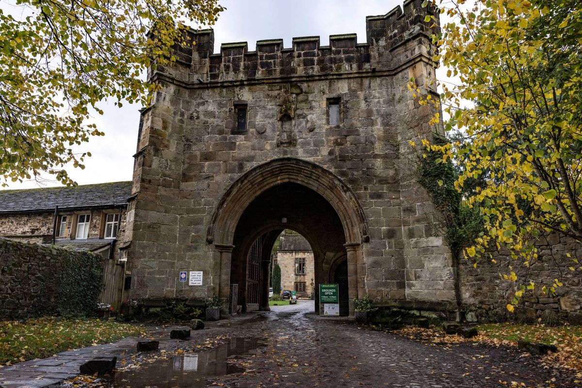 An image depicting an ancient stone archway with crenellations, flanked by lush greenery and autumn foliage, leading into a historical courtyard.