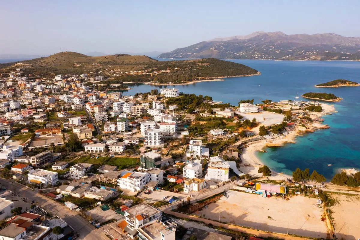An aerial view of a coastal town featuring numerous buildings, a sandy beach, and the expansive blue waters of the sea, with distant hills in the background.