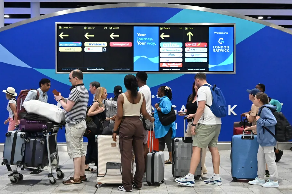 Passengers wait with luggage in the check-in area of Gatwick Airport under a large blue sign with flight information for various airlines.