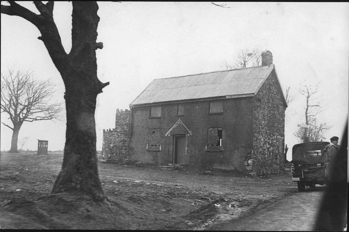 An old black-and-white photograph depicts a solitary, traditional house with a gable roof situated in a rural landscape. A large tree stands on the left side of the image, while a vehicle appears to be driving on a dirt road in the foreground. The scene conveys a sense of historical ambiance with minimal vegetation.