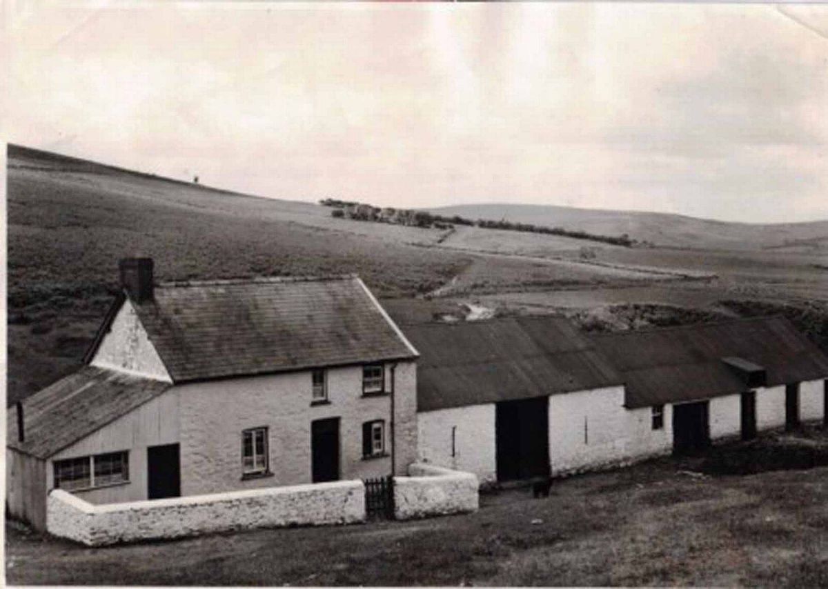 An old black and white photograph depicts a series of buildings, including a central house with a chimney, surrounded by additional structures. The scene is set against a backdrop of rolling hills.