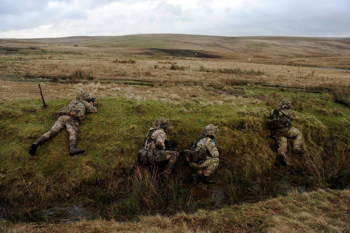 Individuals dressed in camouflage uniforms are engaged in a tactical exercise, utilizing the terrain for strategic advantage. One individual can be seen leaping over a trench, while others are positioned at the edge, adopting defensive postures. The exercise takes place in a vast, open field characterized by sparse vegetation and undulating terrain.