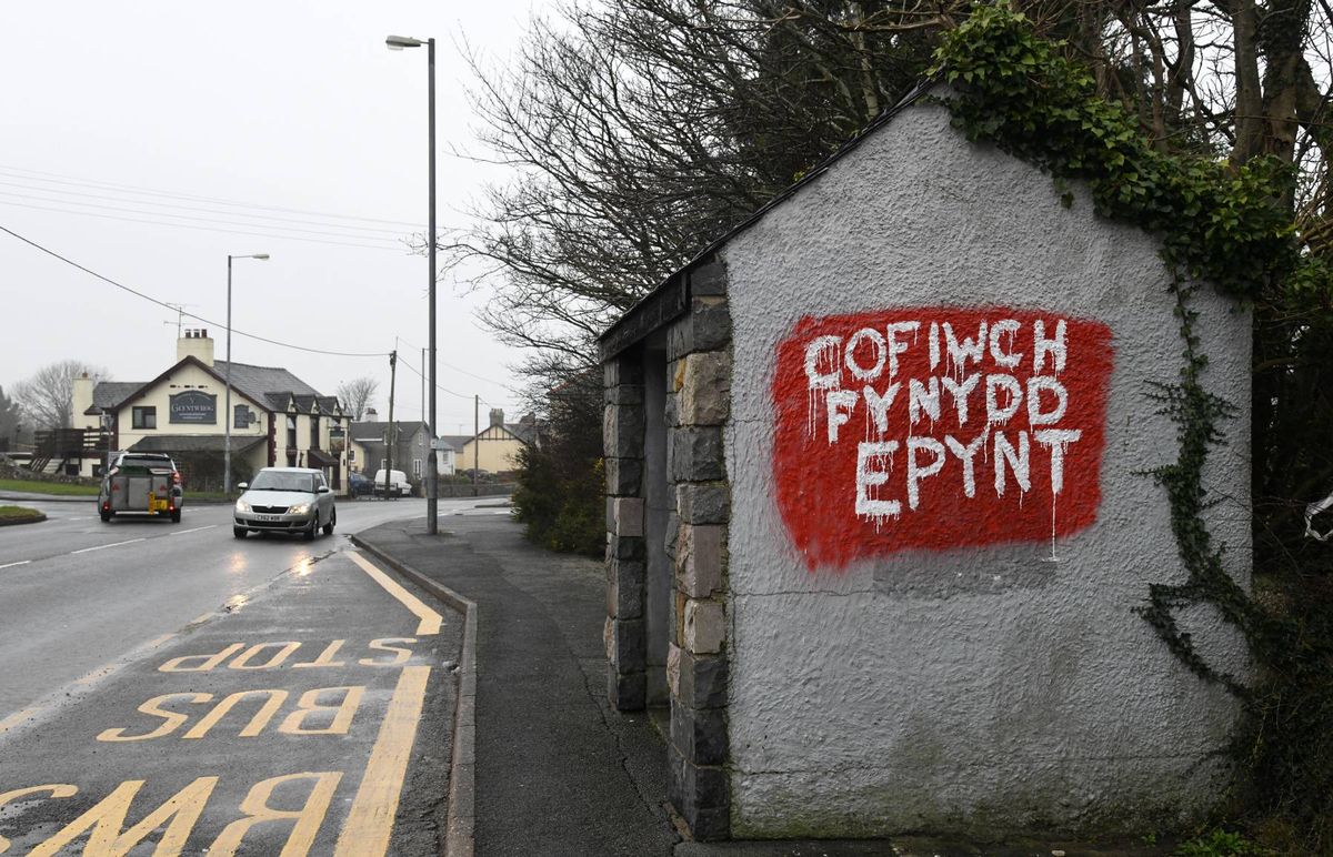 A small building with graffiti on the side, displaying the phrase "GOF FICH PENYDD EPINT" in red letters. The scene is set on a street with a car driving by and a streetlight in the background.