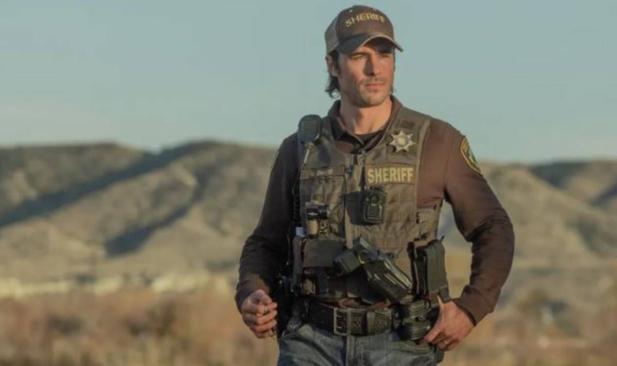 An individual dressed in tactical gear, including a vest and hat, is standing in an open, arid landscape with rugged mountains in the background. The person has their hands on their hips and is holding a firearm.