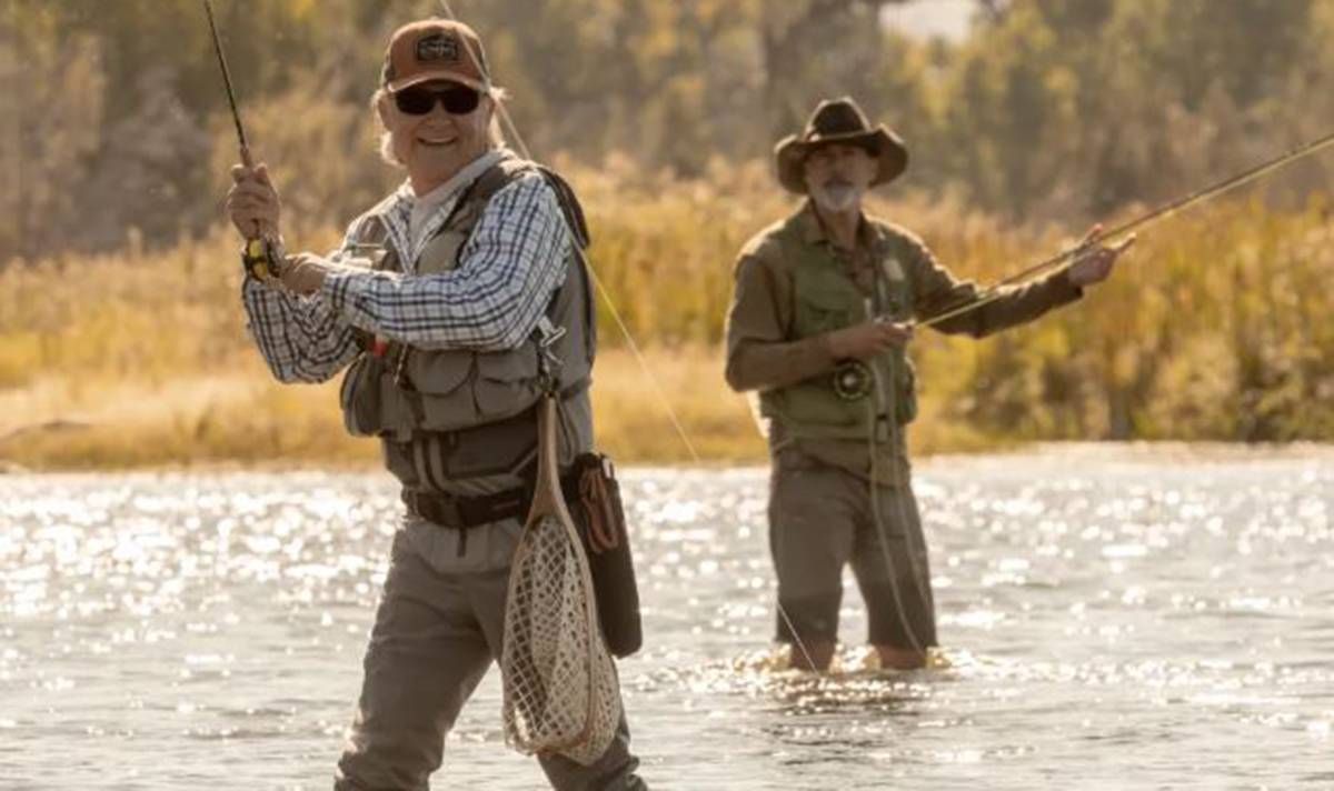 Two individuals engaged in fly fishing, one holding a fishing rod while the other stands nearby, in a serene outdoor setting with a backdrop of tall grass and trees.