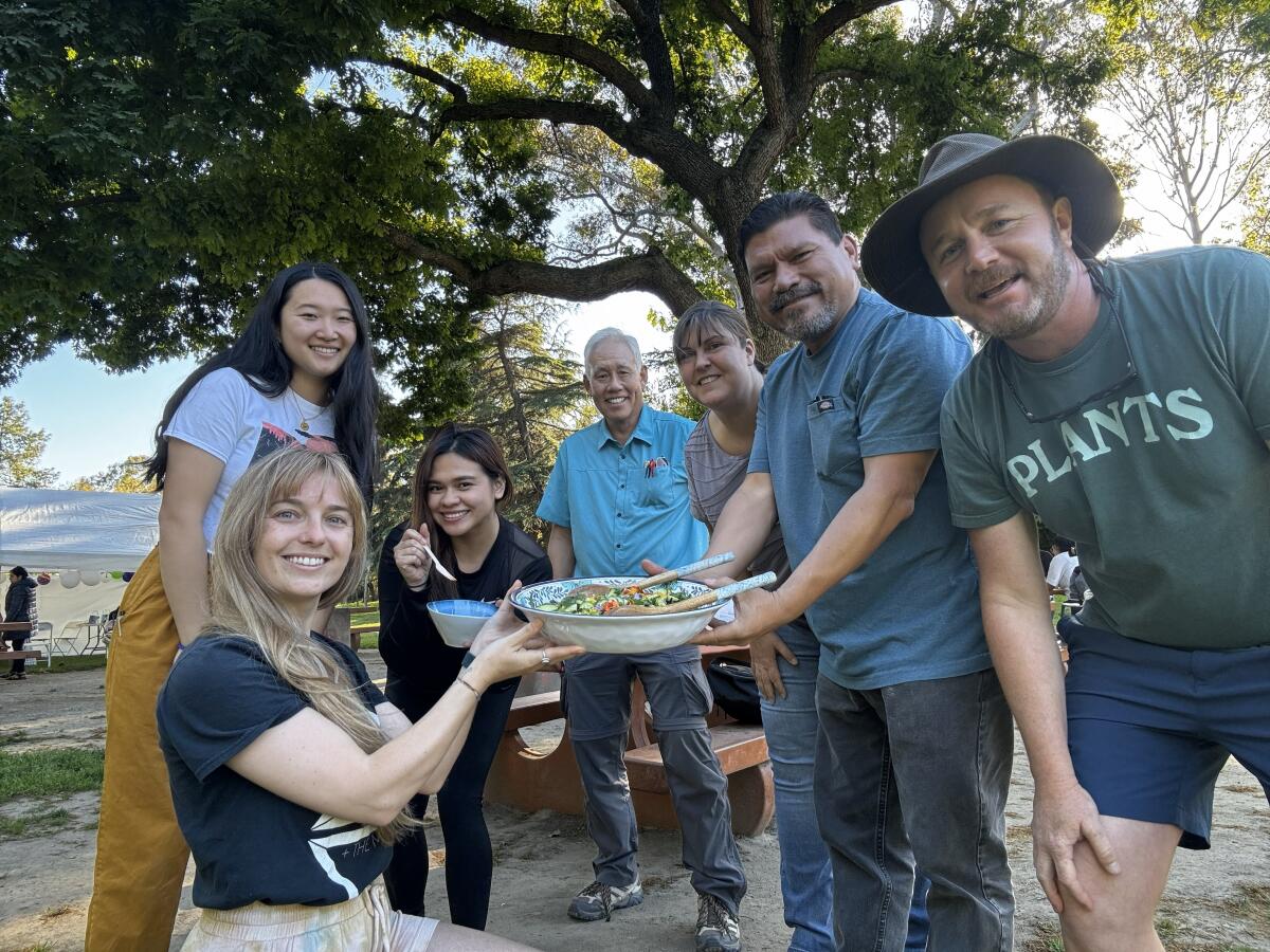 A group of smiling people hold up a large salad bowl full of edible plants.