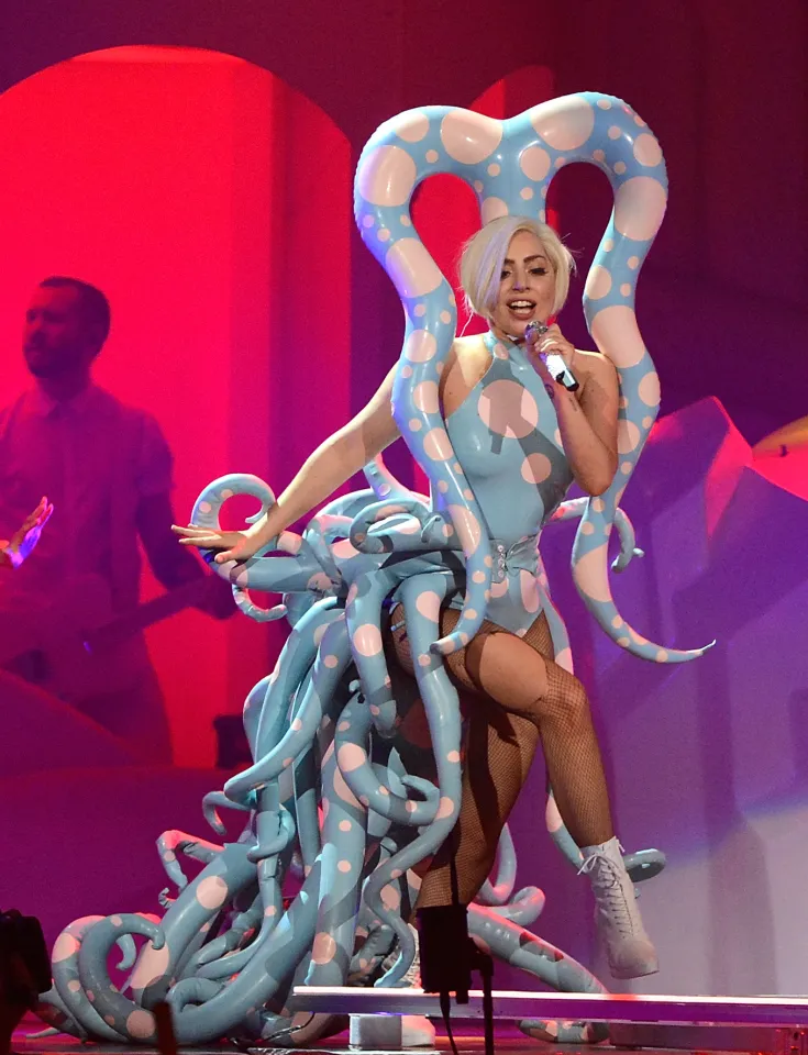 Lady Gaga performs onstage in a light blue polka-dotted octopus-like costume, holding a microphone.