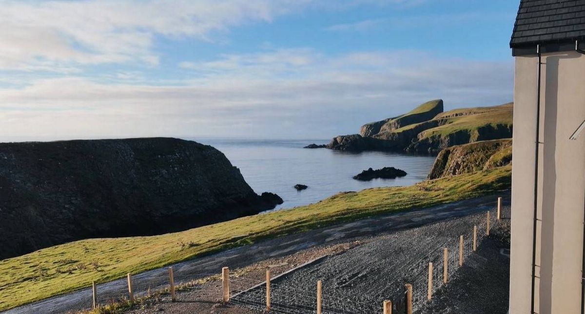Panoramic view of a coastal landscape featuring a rugged cliff with a serene body of water in the foreground, a partially constructed road, and a grassy area adjacent to the shoreline. The sky is clear with a few clouds.