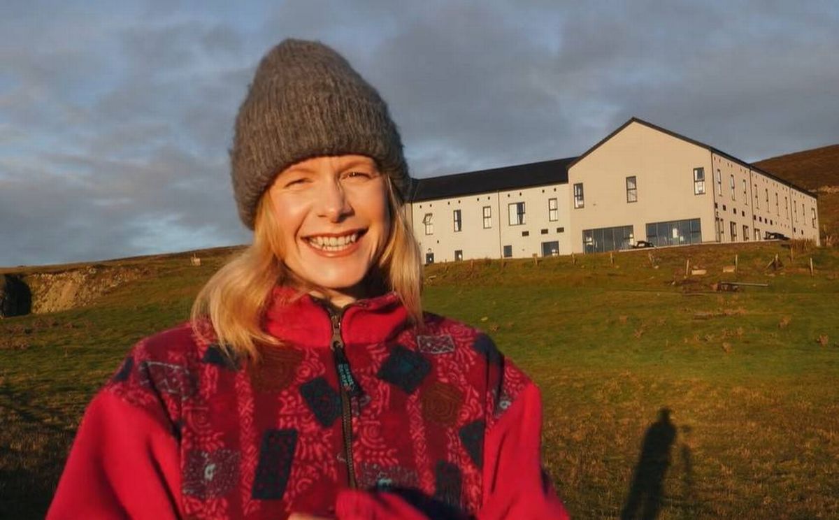 An individual dressed in a red jacket and a beanie poses for a photograph in front of a modern building on a grassy field under a cloudy sky.