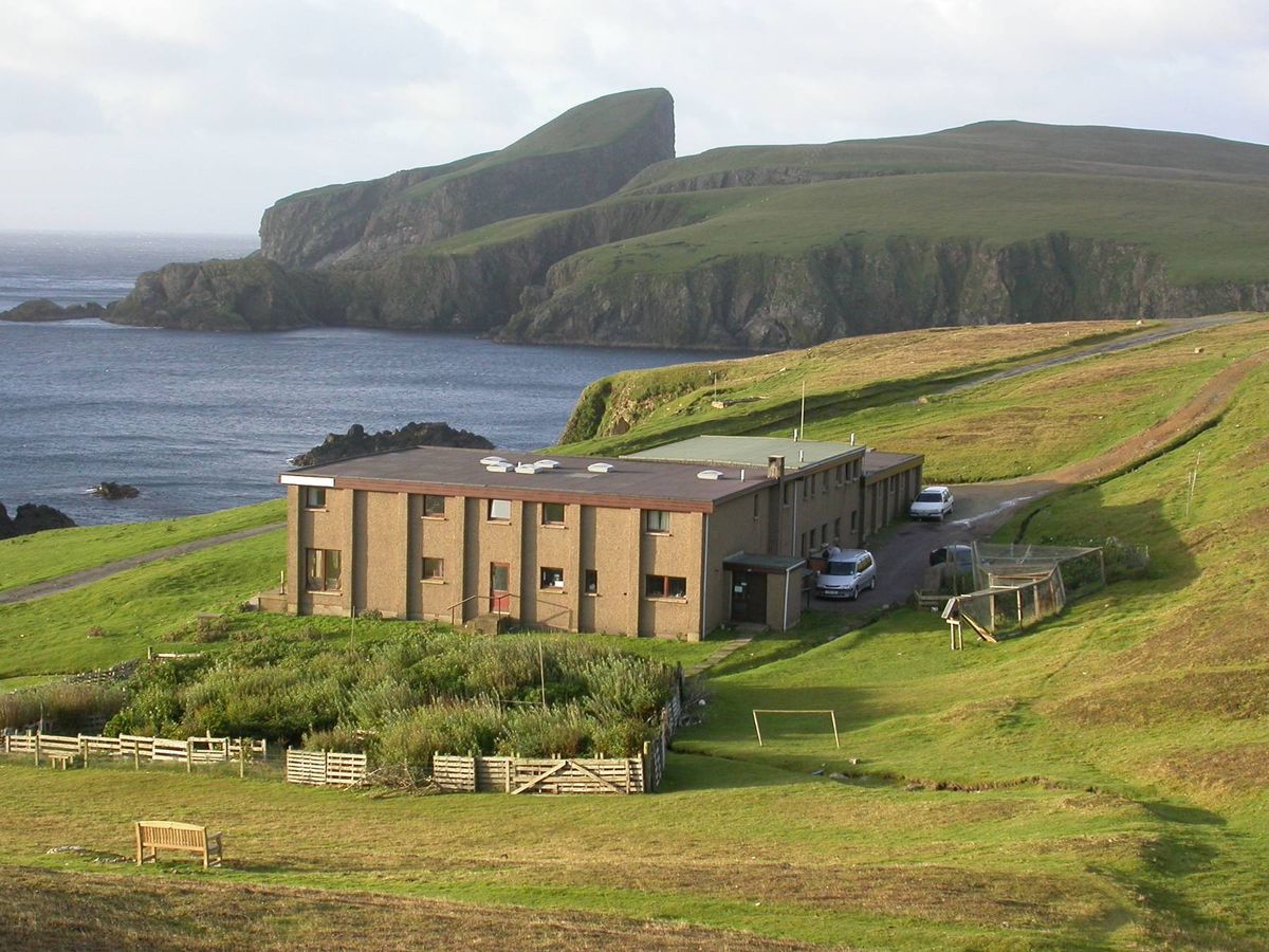 An aerial view of a building situated on a grassy hillside with the sea and a mountainous coastline in the background.