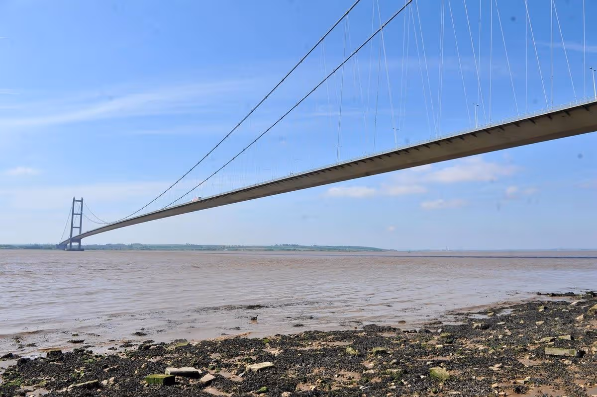 Aerial view of a suspension bridge spanning a body of water, with rocks lining the shoreline and a clear blue sky in the background.