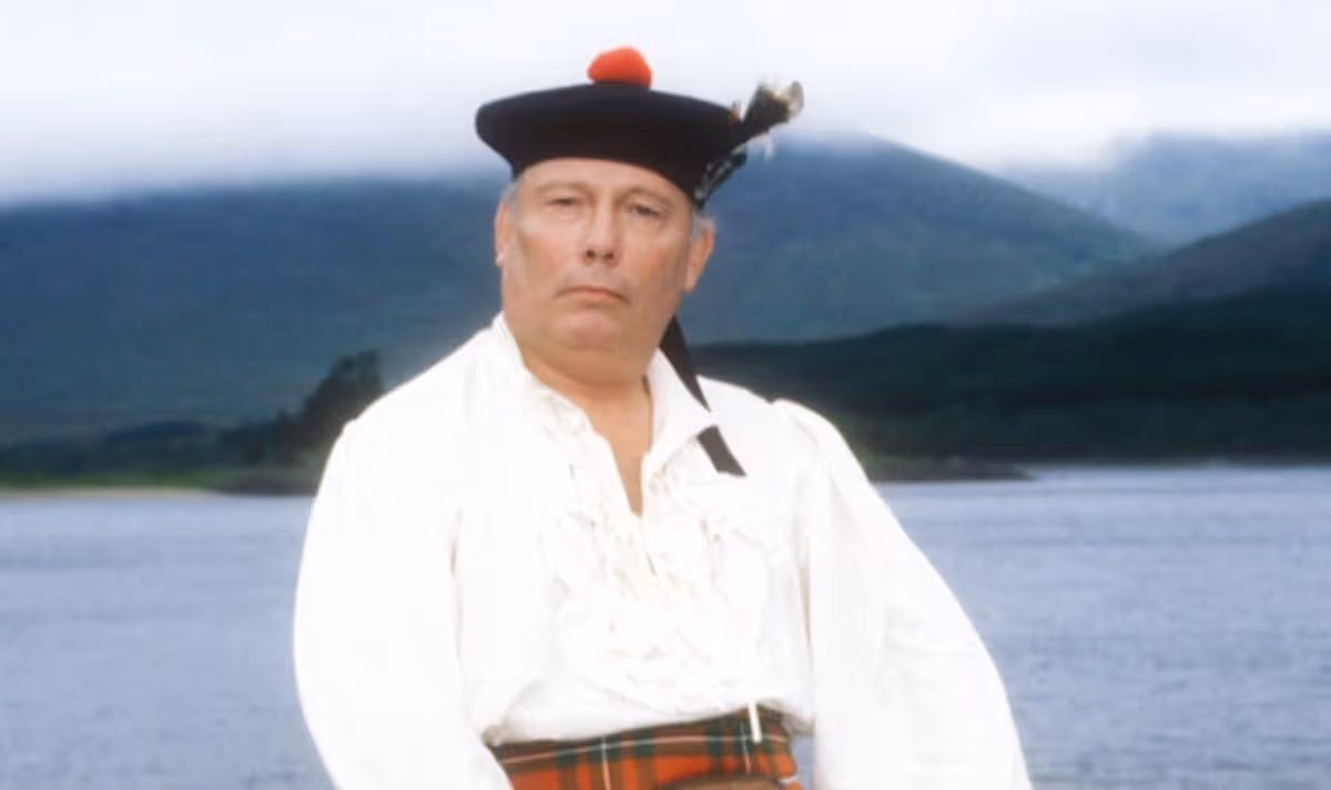A man in a hat and tartan sits by water