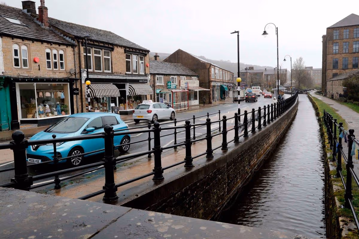 An urban street scene with a canal running alongside, featuring buildings, parked vehicles, and a pedestrian bridge. The canal is partially submerged, and the sky appears overcast.