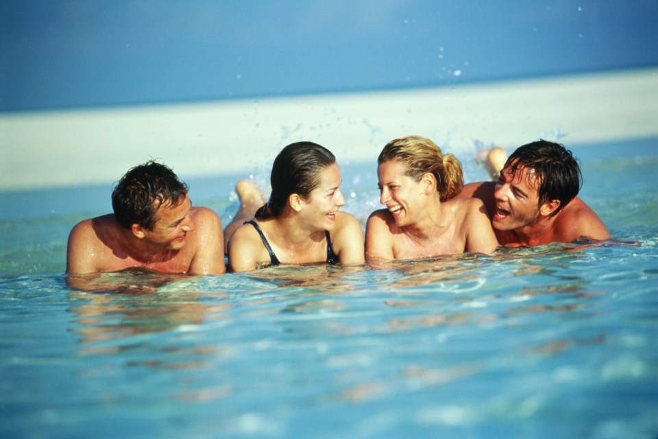 Two couples laughing while lying in shallow water in a swimming pool.