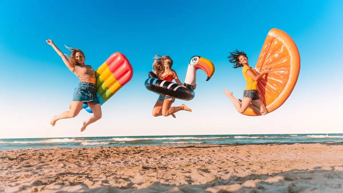 Three happy girls on jumping motion with inflatable mattresses at the beach for celebrating summer vacation - Friends women having fun outdoors on seaside holidays - Joy, carefree and party concept