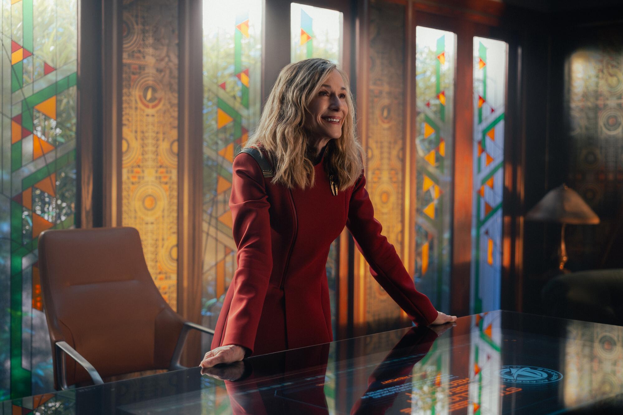 A woman in a red uniform stands and holds both hands on a desk.