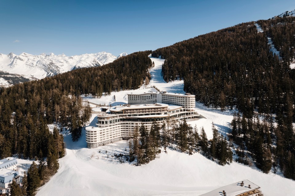 Aerial view of a large, modern white resort hotel built into a snowy mountain ski slope, surrounded by pine trees with snow-capped mountains in the background.
