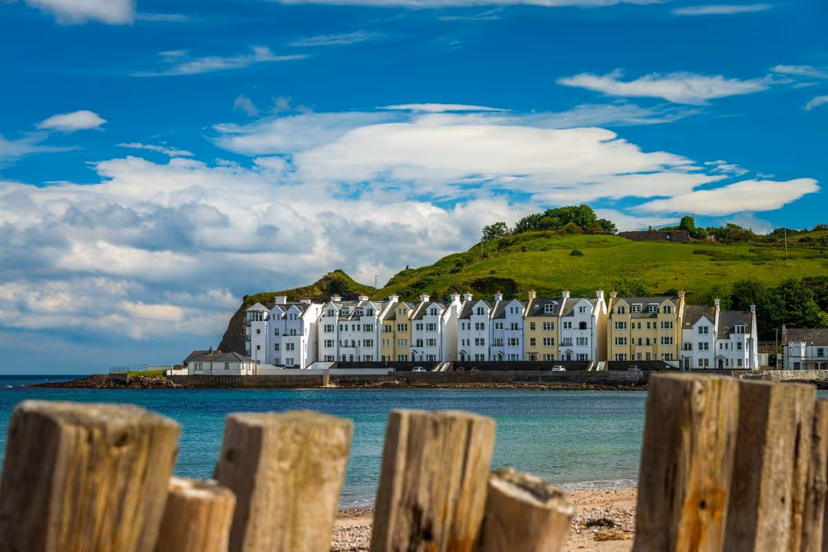 An image of a coastal town, with a large building complex situated on the shoreline, is seen under a blue sky with scattered clouds. The foreground features a wooden barrier, partially obstructing the view of the beach and the ocean.